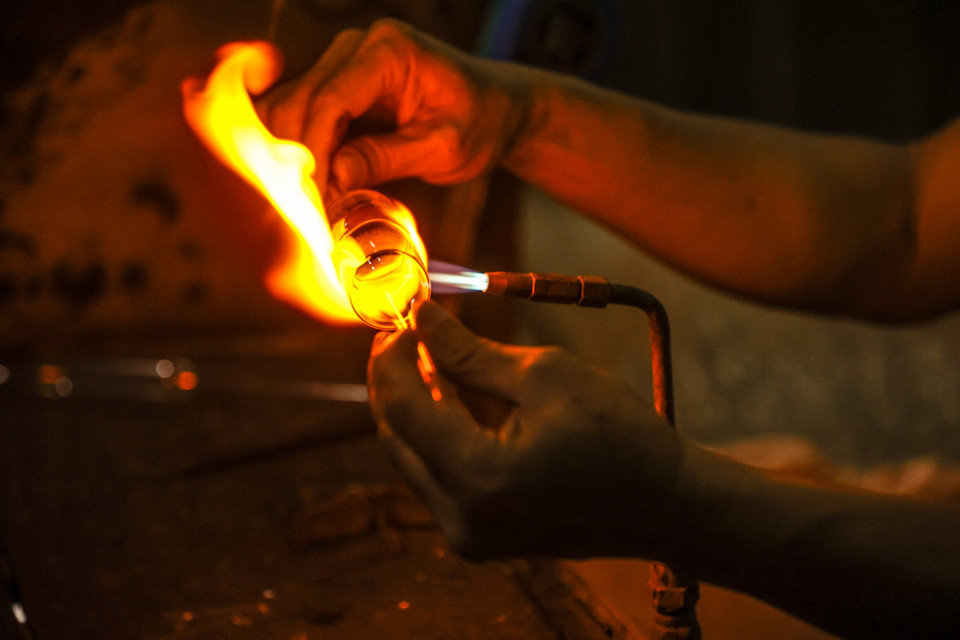 Khi nung nóng thủy tinh trên lửa tới độ 'chín,' người thợ sẽ sử dụng một ống sắt để quết thủy tinh nóng chảy vào đầu ống, sau đó dùng hơi để thổi vào thủy tinh nở phình ra. (Ảnh: Minh Sơn/Vietnam+)