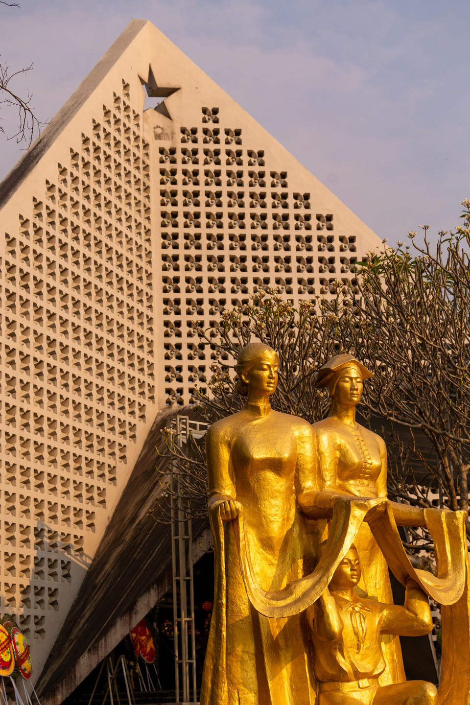 Trước nhà tưởng niệm là hai cây đại thụ và hai cụm tượng màu vàng: một là hai người phụ nữ dân tộc Thái, Kinh và em bé đang nâng dải lụa, hai là hai chiến sỹ mặc áo trấn thủ đội mũ tai bèo. (Ảnh: Minh Sơn/Vietnam+)