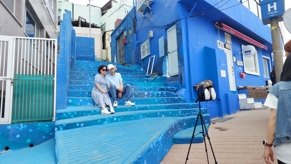 Hai vợ chồng du khách trung niên tự cài đặt máy ảnh ở chế độ tự động và chụp ảnh cùng nhau trong một góc nhỏ màu xanh dương dịu mát. Trong tiếng Hàn, 'huinn' nghĩa là màu trắng, 'yeoul' là lớp sóng nhấp nhô. Trong quá trình an cư lập nghiệp, người dân bản địa cải tạo nhà cửa theo các cung đường núi uốn lượn tựa như nếp sóng và phần lớn được sơn màu trắng, màu xanh dương. (Ảnh: Minh Thu/Vietnam+)