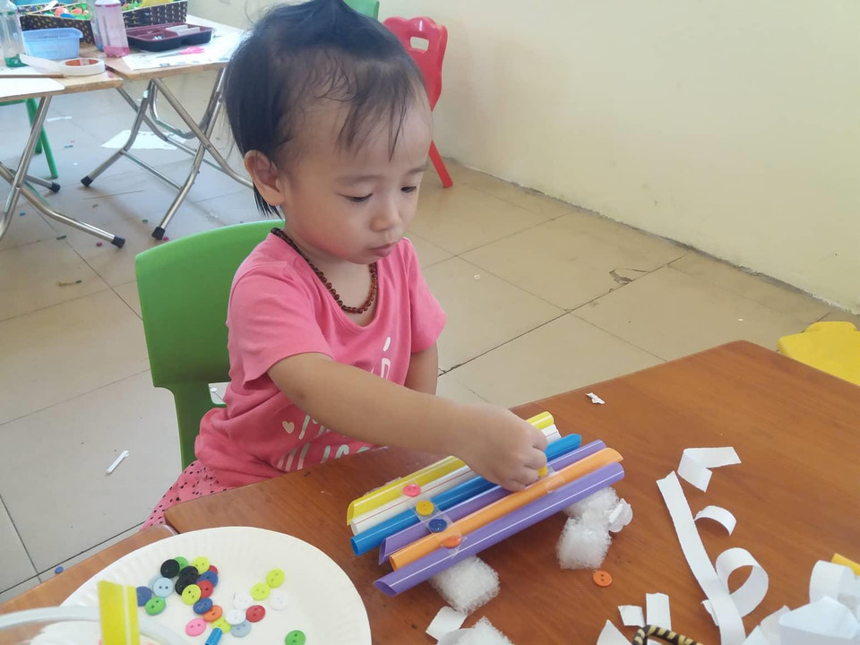 Bé Gia Hân mới hai tuổi, nhưng có rất nhiều hoạt động STEM dành cho lứa tuổi này. Trong ảnh, Gia Hân đang dùng cúc áo nhiều màu để trang trí chiếc thuyền làm từ ống hút. (Ảnh: PV/Vietnam+)