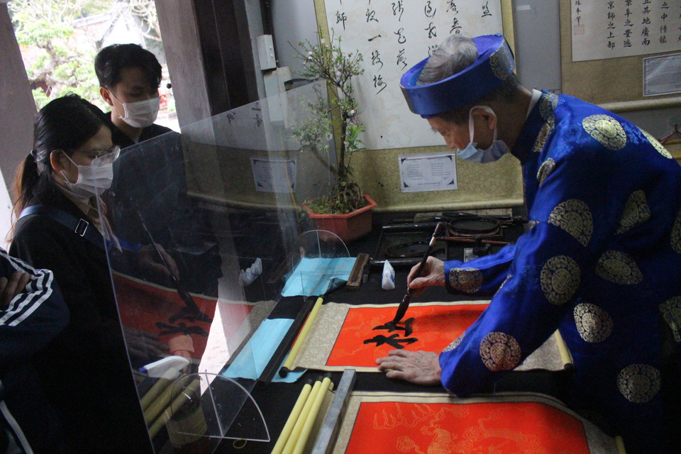 Hoạt động xin chữ ở đây cũng đã trở lại nhưng chỉ có một quầy. Ông đồ viết thư pháp sau kính chắn giọt bắn và tuân thủ quy định đeo khẩu trang liên tục khi "cho chữ". (Ảnh: Minh Anh/Vietnam+)