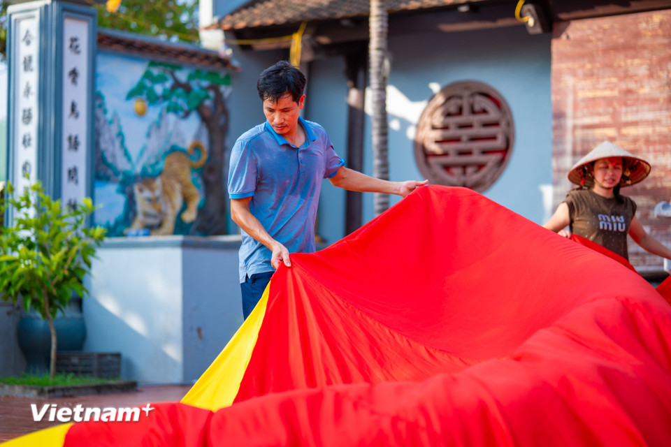 Để thực hiện một lá cờ có kích thước tương 30m x 20m, anh Trung phải huy động tổng lực người làm. Trước tiên, anh Trung kiểm tra dự báo thời tiết, chỉ khi trời nắng ráo, không mưa, họ mới trải vải lớn trên mặt bằng phẳng đủ rộng thường là sân đình làng để ghép các khổ vải lại với nhau. (Ảnh: Minh Sơn/Vietnam+)