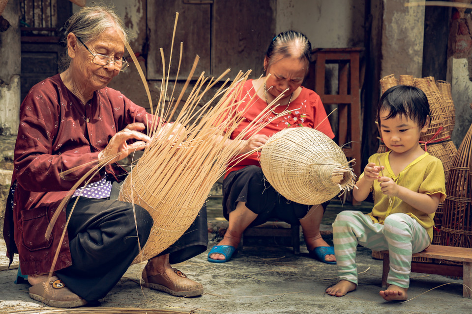 Những người cao tuổi đôi tay vẫn thoăn thoắt đường nan, vừa làm vừa kể chuyện cho lớp trẻ, tạo môi trường tiếp xúc nghề từ khi còn tấm bé. (Ảnh: Hoài Nam/Vietnam+)