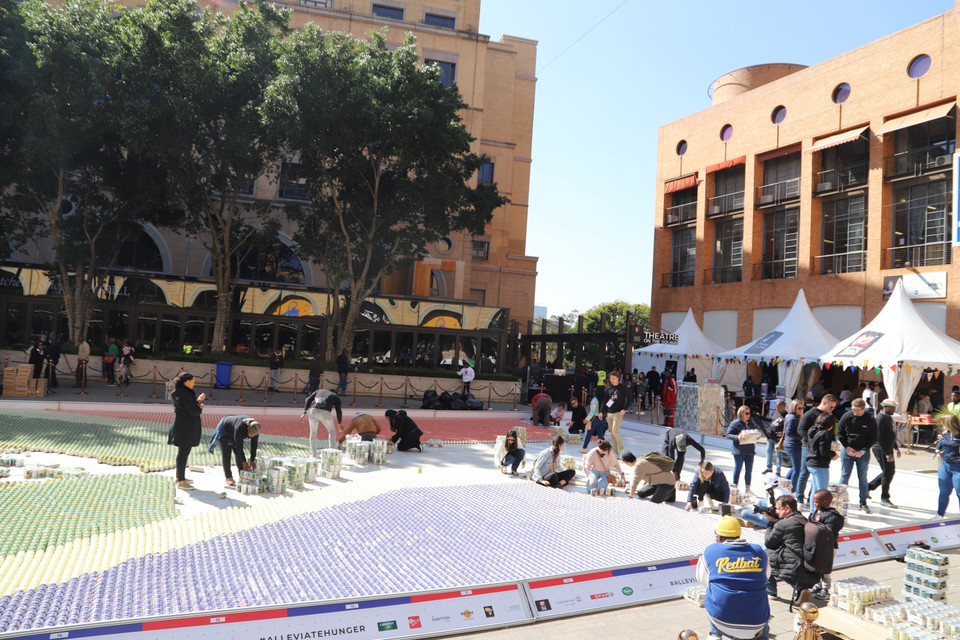 Với sự ủng hộ của các nhà tài trợ doanh nghiệp thực phẩm và đồ uống cùng sự hỗ trợ của các tình nguyện viên nhiệt tình, tổ chức từ thiện này đã tổ chức việc sắp đặt gần 90.000 lon thực phẩm trên hai bức tranh ghép khổng lồ ở Trung tâm hội nghị quốc tế ở thành phố Cape Town và Quảng trường Nelson Mandela tại thành phố Johannesburg trong cùng một ngày. (Ảnh: Hồng Minh/TTXVN) 