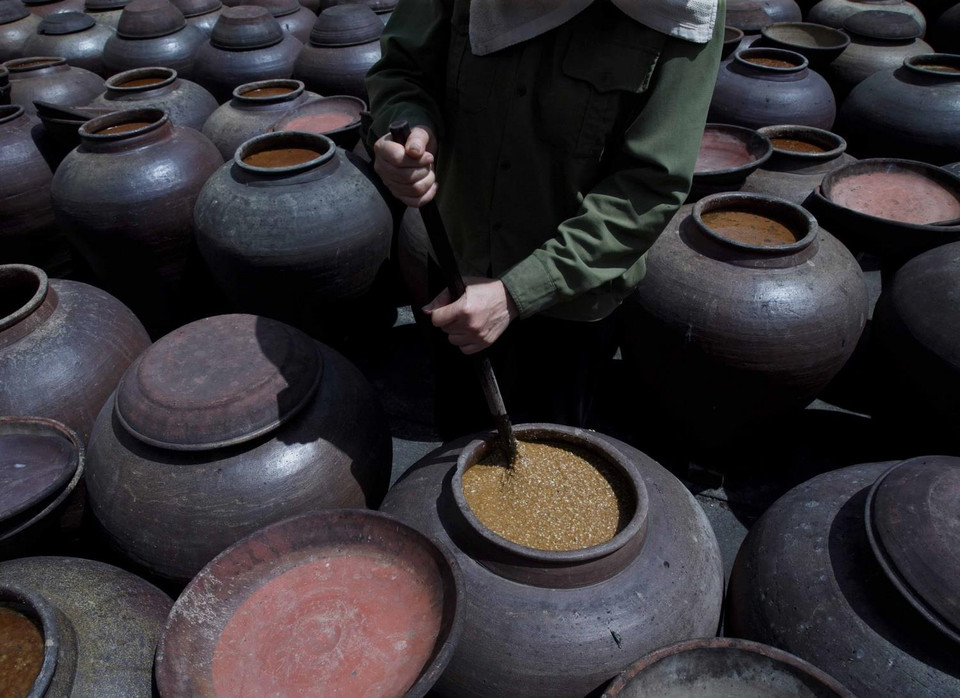 Để tương nhuyễn và lên màu, người làm tương phải thường xuyên khuấy tương vào buổi sáng và trưa. Ánh sáng mặt trời làm nóng chum, giúp tương chín nhanh hơn. Trời càng nhiều nắng thì tương chín càng nhanh. (Ảnh: Phạm Kiên/TTXVN)