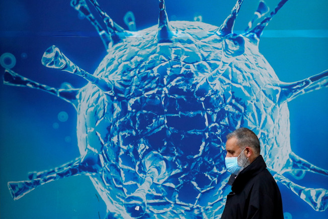 Người dân đeo khẩu trang đi ngang qua hình ảnh minh họa cho virus SARS-CoV-2. (Ảnh: Reuters) Biến thể phụ BA.2 có khả năng lây nhanh gấp 1,5 lần Omicron ảnh 1