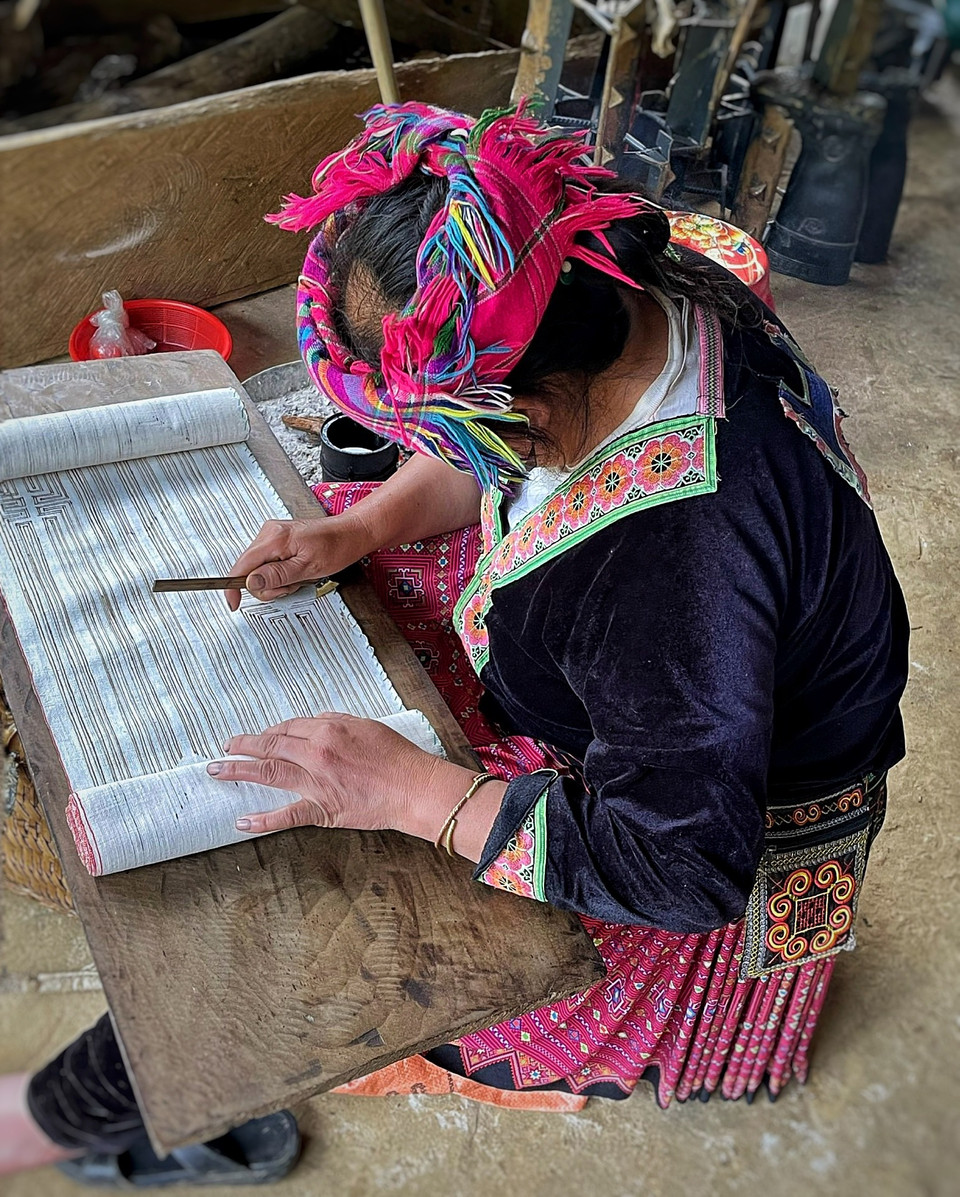 Để vẽ được sáp ong lên vải, phụ nữ Mông có một bộ bút vẽ làm bằng thanh tre nhỏ, ngòi bút là một lá đồng nhỏ hình tam giác được nẹp vào thanh tre. Khi vẽ luôn phải ngồi bên bếp lửa, chấm bút vào chảo sáp ong nóng đặt trên than hồng rồi thoăn thoắt đưa tay kẻ những đường thẳng trên vải, sao cho lưỡi bút song song với mặt đất. Phải kẻ thật đều thật khéo để lượng sáp chảy đều, không loang lổ cho đến hết rồi mới tiếp tục chấm tiếp.(Ảnh: Xuân Mai/Vietnam+)