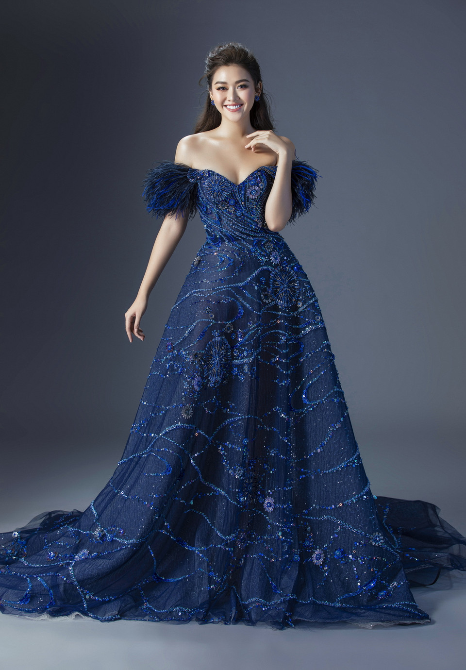  Được biết hai bộ dạ hội này đều của nhà thiết kế Đỗ Long. Bộ dạ hội xanh nổi bật với chi tiết đính cườm, thiết kế đầm trễ vai khoét sâu phần ngực khoe đôi vai trần quyến rũ. Đặc biệt, chi tiết lông vũ hai bên cầu vai giúp tôn lên vẻ mềm mại, nét đẹp hiền hậu và không kém phần sang trọng. (Ảnh: Lê Thiện Viễn/Vietnam+)