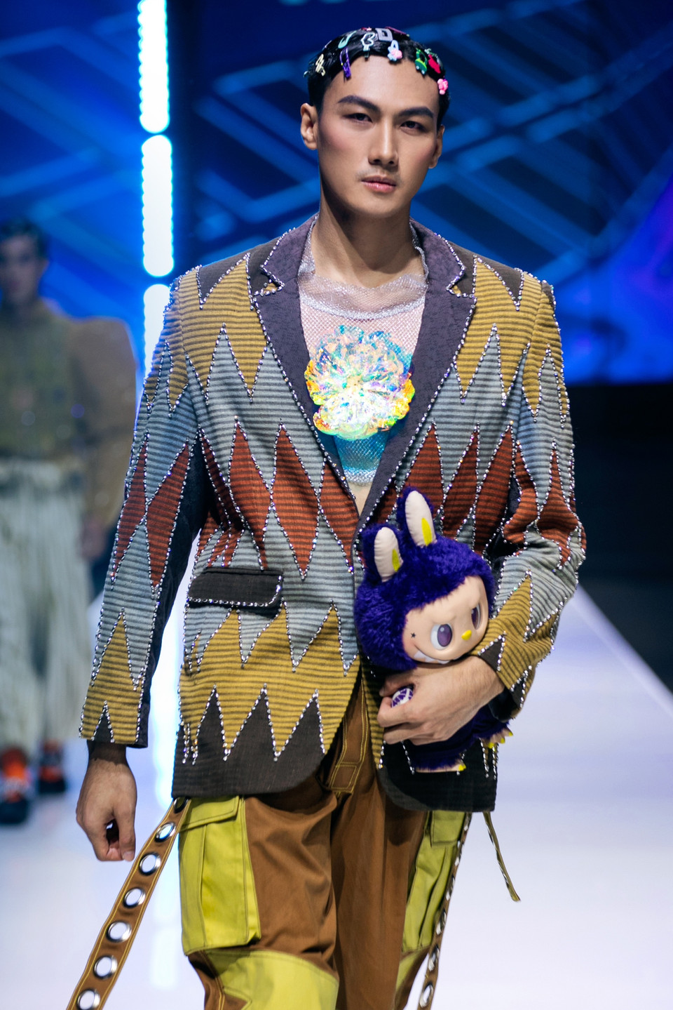 Đảm nhiệm kết màn show diễn, quán quân The Face Vietnam Mạc Trung Kiên diện trang phục có họa tiết zig-zag cùng màu sắc nổi bật, kết hợp quần nâu rộng với khoen kim loại hai bên, tạo phong cách mạnh mẽ. Phụ kiện cài tóc và giày ánh bạc cùng gấu bông Labubu tô đậm phong thái trẻ trung. (Ảnh: PV/Vietnam+)