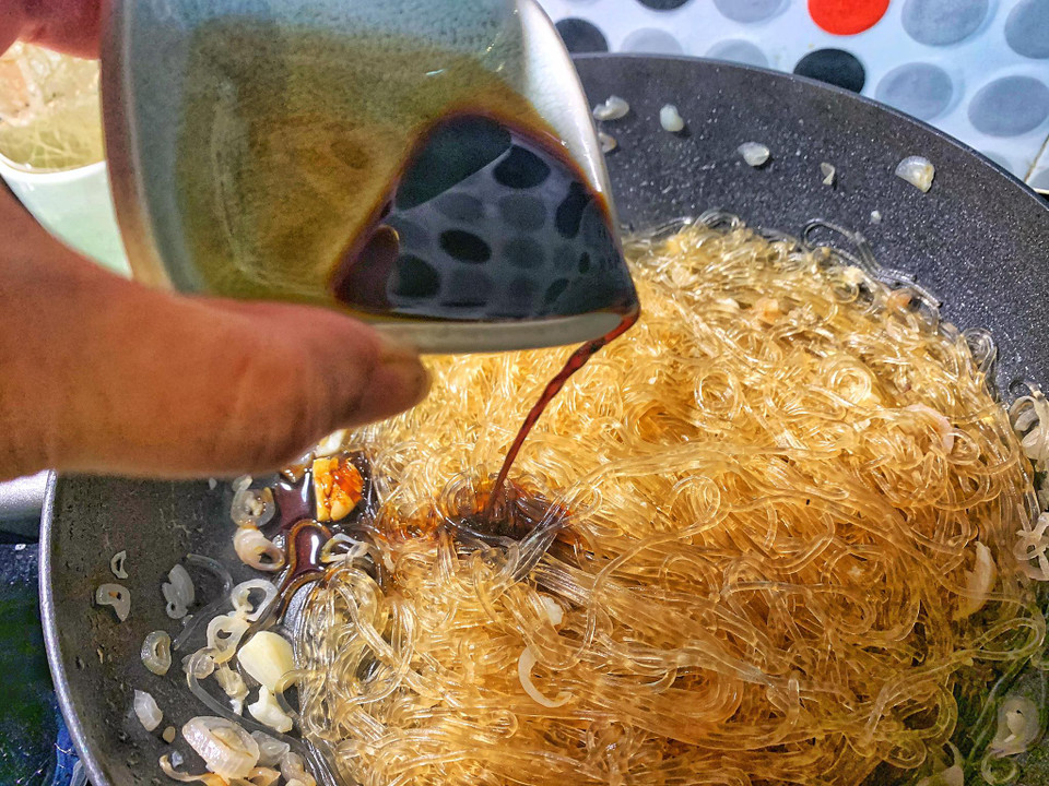 Phi thơm số hành, tỏi còn lại để xào miến. Bước này là lúc bạn cho bát nước xương hay nước luộc gà vào để miến không bị khô và 'ngậm' chất béo từ nước xương. Để màu miến đẹp, cho 2 thìa xì dầu càng cua vào đảo đều. (Ảnh: Mai Mai/Vietnam+)