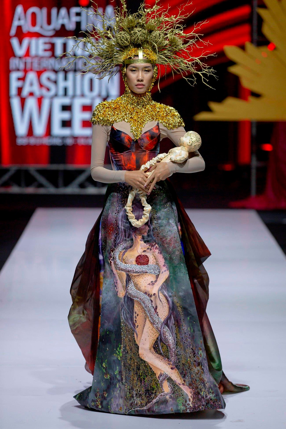 Khoác lên mình bộ trang phục được in màu hài hoà với sự phân bố các layers họa tiết đa sắc trên nền xanh, đan xen yếu tố tôn giáo, người mẫu Nguyễn Oanh bế trên tay đứa bé vừa được 'thoát thai' đúng nghĩa, mang đến cảm giác vừa thích thú lại vừa kỳ dị cho khán giả. (Ảnh: CTV)