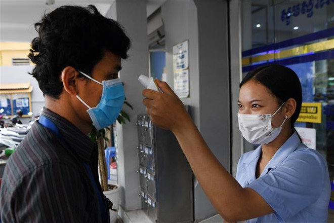 Nhân viên y tế kiểm tra thân nhiệt một khách hàng nhằm ngăn dịch COVID-19 lây lan, bên ngoài một ngân hàng ở Phnom Penh, Campuchia, ngày 17/3/2020. (Ảnh: AFP/TTXVN) Cập nhật tình hình sức khỏe của 13 người Việt mắc COVID-19 ở Campuchia ảnh 1