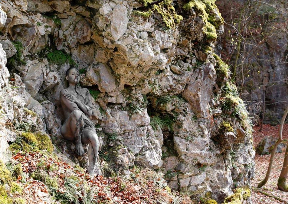 Người mẫu ngồi phía trước mỏm đá với cơ thể được sơn màu xanh lá và màu xám.