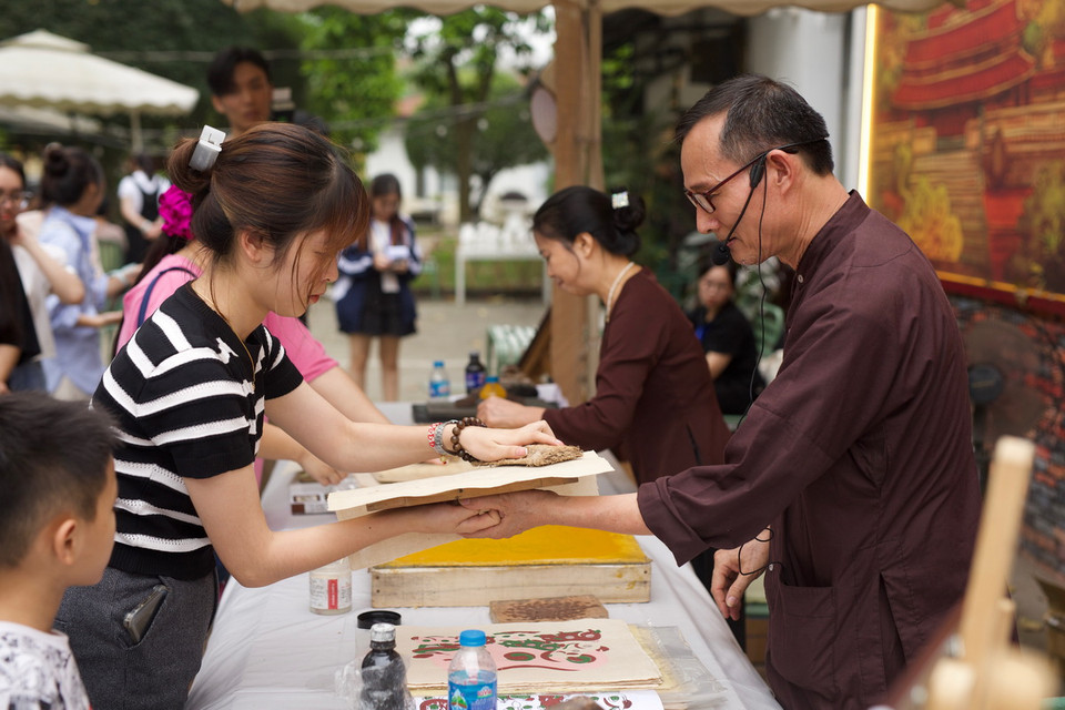 Bước đầu tiên, sau khi quét nền, dùng thét quét màu vàng lên bìa, sau đó rập ván in lên bìa vài lần, đặt ván in lên giấy theo cữ, tay trái luồn xuống dưới tờ giấy lật cả giấy lẫn ván lên rồi dùng xơ mướp xoa lên phía sau tờ giấy để màu bắt đều nét, cuối cùng bóc tờ tranh ra bỏ sang bên trái, các tờ in sau để so le với tờ trước. (Ảnh: Đặng Yến/Vietnam+)