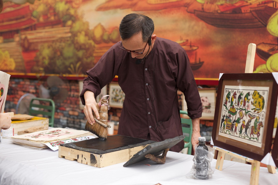 Bước đầu tiên, sau khi quét nền, dùng thét quét màu vàng lên bìa, sau đó rập ván in lên bìa vài lần, đặt ván in lên giấy theo cữ, tay trái luồn xuống dưới tờ giấy lật cả giấy lẫn ván lên rồi dùng xơ mướp xoa lên phía sau tờ giấy để màu bắt đều nét, cuối cùng bóc tờ tranh ra bỏ sang bên trái, các tờ in sau để so le với tờ trước. (Ảnh: Đặng Yến/Vietnam+)