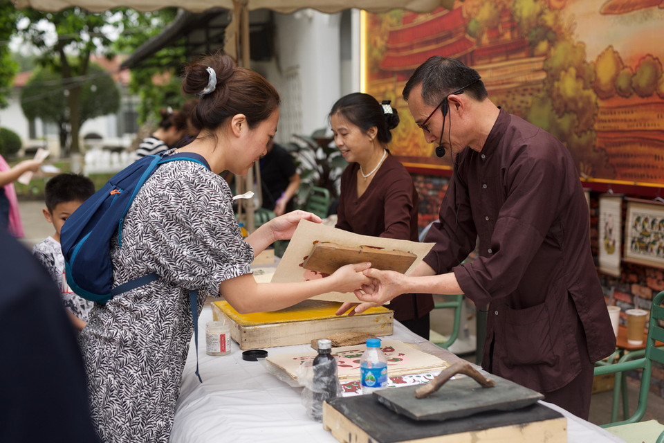 Bước đầu tiên, sau khi quét nền, dùng thét quét màu vàng lên bìa, sau đó rập ván in lên bìa vài lần, đặt ván in lên giấy theo cữ, tay trái luồn xuống dưới tờ giấy lật cả giấy lẫn ván lên rồi dùng xơ mướp xoa lên phía sau tờ giấy để màu bắt đều nét, cuối cùng bóc tờ tranh ra bỏ sang bên trái, các tờ in sau để so le với tờ trước. (Ảnh: Đặng Yến/Vietnam+)