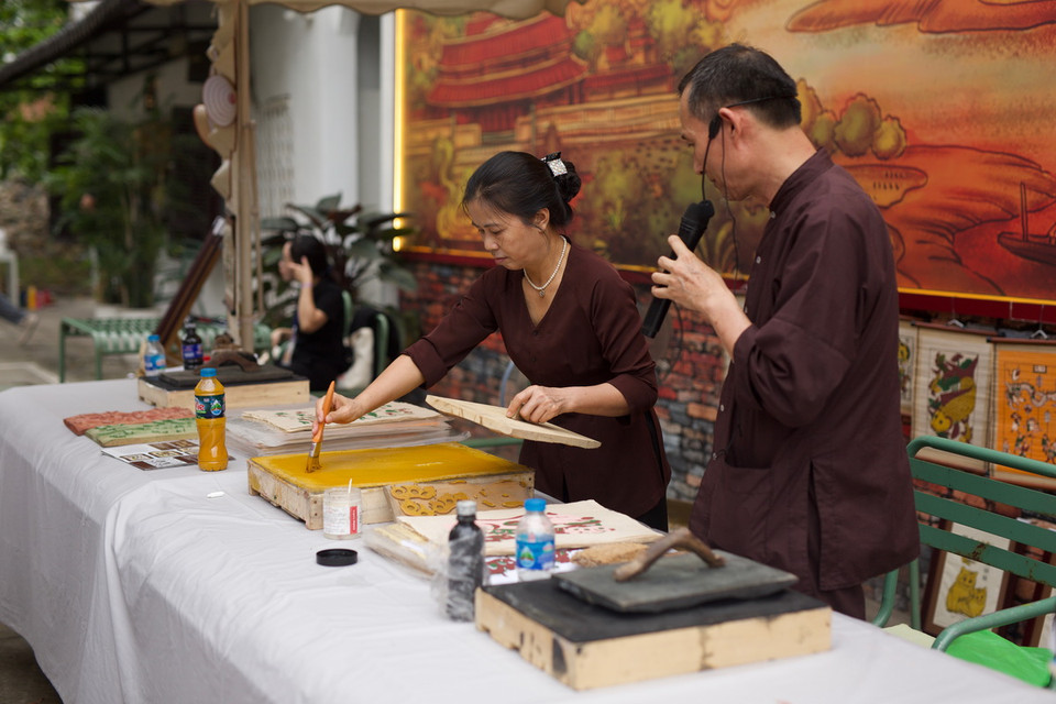 Nguyên vật liệu in tranh bao gồm: bản cắt, giấy dó, điệp, thét, bìa, xơ mướp. Bước đầu tiên, sau khi quét nền, dùng thét quét màu vàng lên bìa, sau đó rập ván in lên bìa vài lần, đặt ván in lên giấy theo cữ, tay trái luồn xuống dưới tờ giấy lật cả giấy lẫn ván lên rồi dùng xơ mướp xoa lên phía sau tờ giấy để màu bắt đều nét, cuối cùng bóc tờ tranh ra bỏ sang bên trái, các tờ in sau để so le với tờ trước. (Ảnh: Đặng Yến/Vietnam+)
