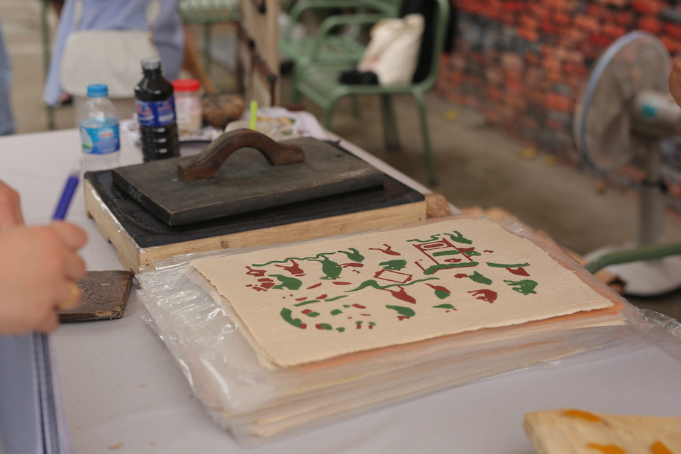 Bước đầu tiên, sau khi quét nền, dùng thét quét màu vàng lên bìa, sau đó rập ván in lên bìa vài lần, đặt ván in lên giấy theo cữ, tay trái luồn xuống dưới tờ giấy lật cả giấy lẫn ván lên rồi dùng xơ mướp xoa lên phía sau tờ giấy để màu bắt đều nét, cuối cùng bóc tờ tranh ra bỏ sang bên trái, các tờ in sau để so le với tờ trước. (Ảnh: Đặng Yến/Vietnam+)