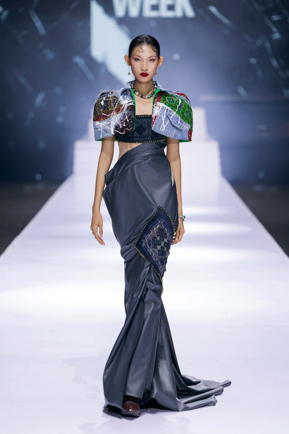 Quán quân The Face Vietnam 2023 Huỳnh Tú Anh mở màn show diễn nhẹ nhàng nhưng đầy cuốn hút, mở lối cho hành trình “mộng du” của bộ sưu tập. Cô diện thiết kế lấy cảm hứng từ dáng váy quấn, bộ trang phục được xử lý bằng chất liệu da bóng với kỹ thuật draping tạo phom sắc sảo. Phần áo khoác crop-top vai vuông phối các mảng denim và thổ cẩm tạo điểm nhấn mạnh mẽ, tôn lên tinh thần văn hóa trong ngôn ngữ thiết kế. (Ảnh: CTV/Vietnam+)