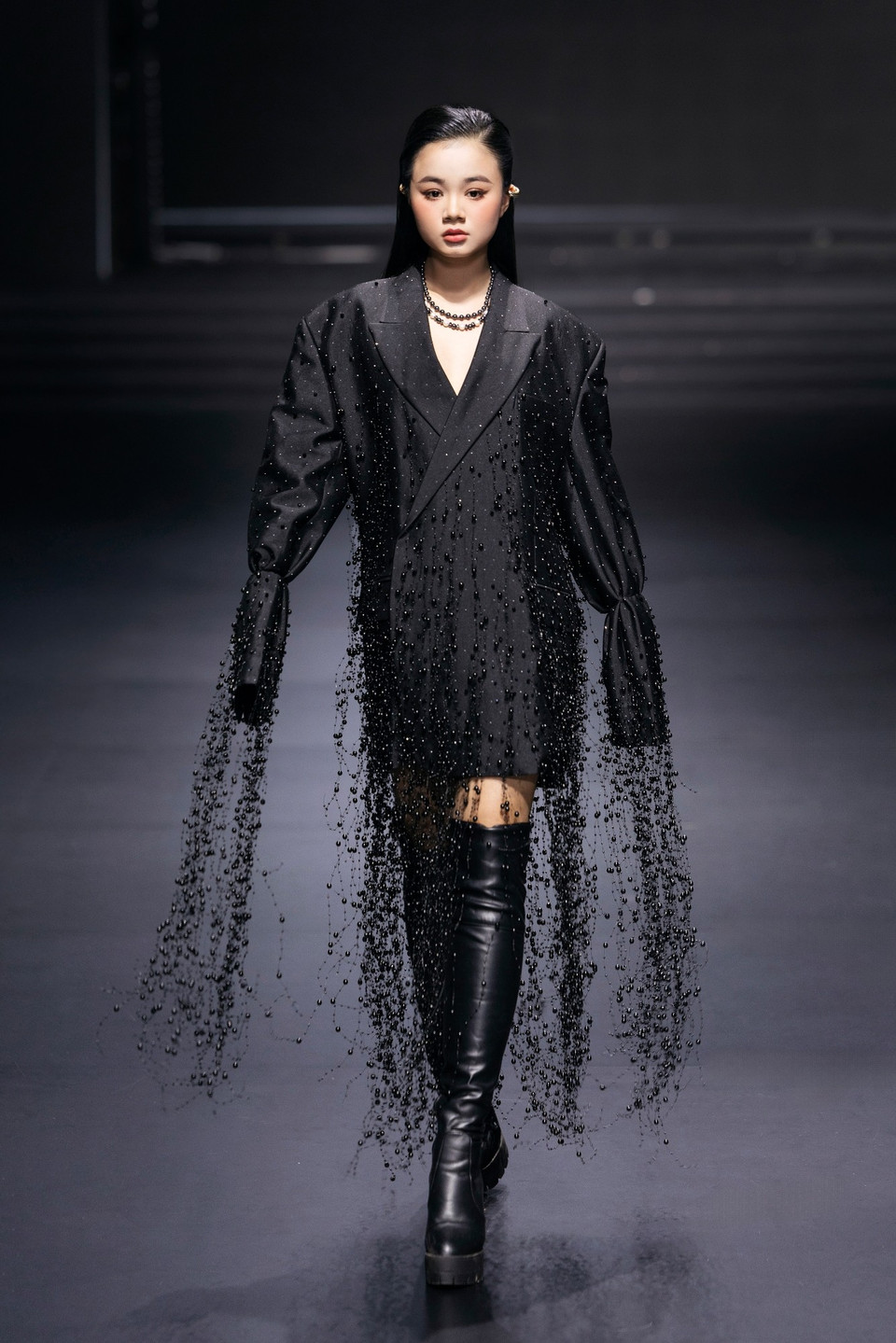 Mở đầu là thiết kế blazer dáng dài cách điệu kết hợp kỹ thuật đính kết thủ công tỉ mỉ bằng hàng trăm viên ngọc trai đen và hạt pha lê ánh kim trải đều từ phần thân áo đến các dải tua rua mềm mại buông dài từ tay áo. Hiệu ứng chuyển động khi người mẫu sải bước tạo cảm giác như những tia nước lấp lánh tung bay trong ánh đèn đêm, đúng với tinh thần “Evening Pool Party” – quyến rũ, phóng khoáng và lấp lánh. (Ảnh: CTV/Vietnam+)