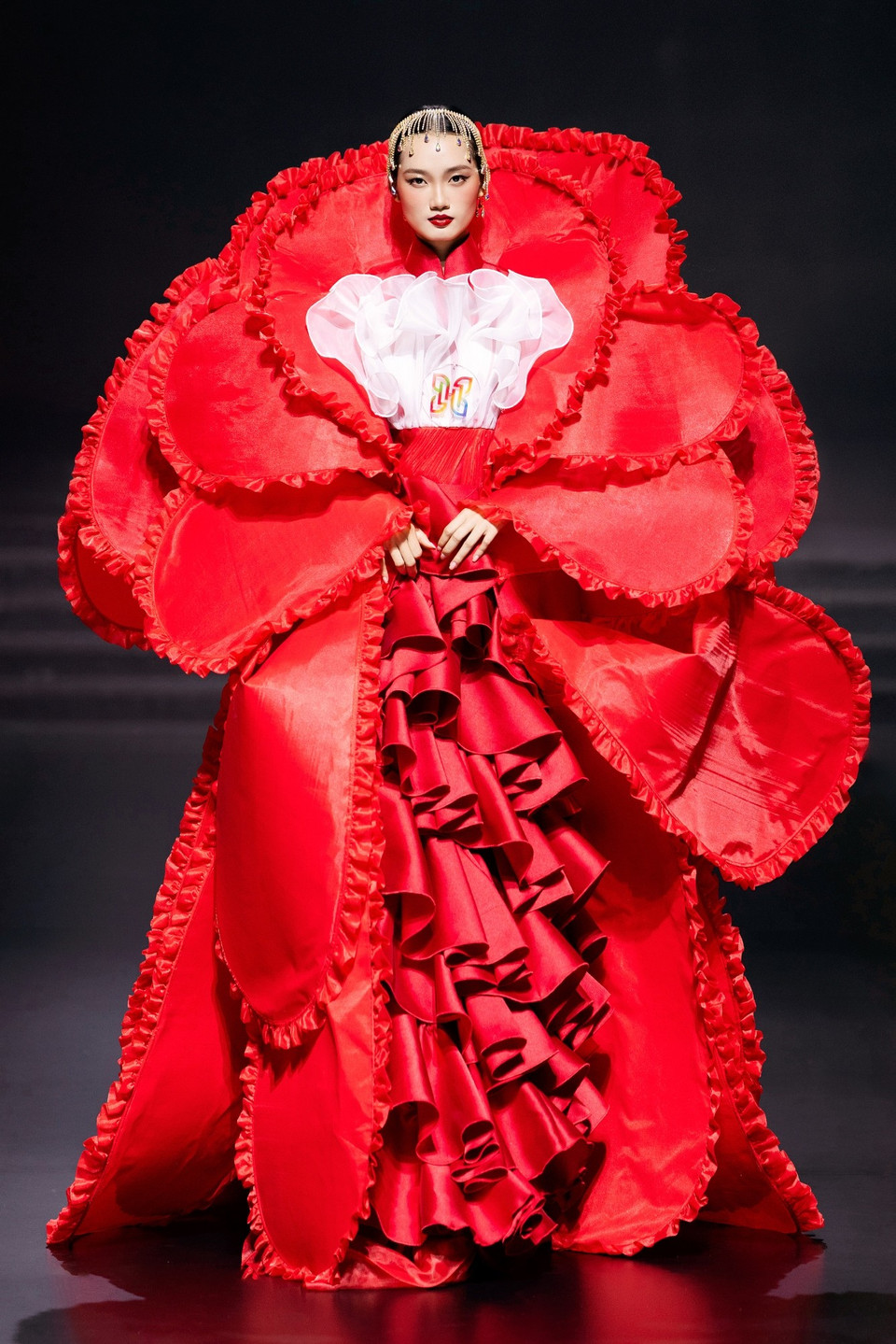 Thiết kế mở màn của bộ sưu tập gây ấn tượng với sự xuất hiện đầy kiêu sa của Á hậu Quỳnh Anh, hóa thân thành một đóa hồng rực rỡ giữa ánh bình minh. Thiết kế sử dụng kỹ thuật xếp tầng và dựng phom 3D tinh xảo để tạo nên những cánh hoa lớn bản, xếp lớp đối xứng bao quanh cơ thể, tái hiện hình ảnh một bông hồng đang bung nở tràn đầy năng lượng. (Ảnh: CTV/Vietnam+)