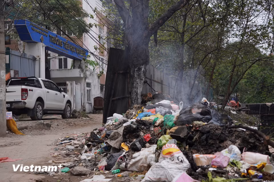 Theo ghi nhận của phóng viên Báo Điện tử VietnamPlus, rất nhiều điểm tập kết rác ngay trước cổng trường Tiểu học Vạn Phúc. Chị Ngô Thị H.L (phụ huynh có con học tại Trường Tiểu học Vạn Phúc) lo lắng: “Mỗi ngày đưa đón con đi học là một cực hình, đứng đợi đón con ở đây là phải nín thở. Mương ngay sát trường học, ruồi muỗi nhiều, mùi hôi thối nồng nặc nên tôi rất lo ngại sẽ ảnh hưởng đến sức khỏe lâu dài của bọn trẻ.” (Ảnh: Vân Anh/Vietnam+)