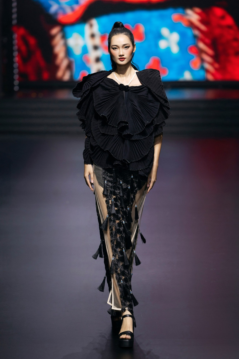 Á hậu Quỳnh Anh mở màn show diễn trong bộ trang phục mang tính tạo hình mạnh mẽ: phần thân trên xếp ly thủ công được đặt để từng tầng cầu kỳ, mô phỏng đường cong mềm mại của sườn núi Tây Bắc, trong khi thân dưới gây ấn tượng với lớp voan trong suốt, điểm tua rua dọc thân mang hiệu ứng chuyển động uyển chuyển theo từng bước catwalk. (Ảnh: CTV/Vietnam+)