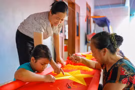 Cháu Huệ thường xuyên tới nhà chị Nhung để thêu cờ cùng các bậc cao niên. Bà Thiết và chị Nhung hướng dẫn bé Huệ đi đường kim sao cho chính xác và đẹp nhất. (Ảnh: Hoài Nam/Vietnam+)