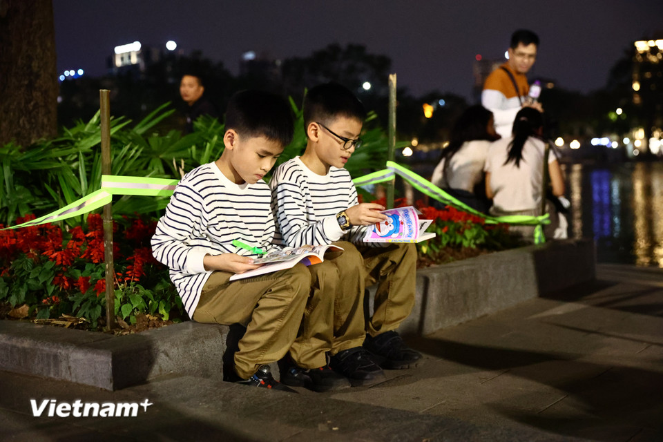 Hai em nhỏ ngồi đọc truyện ven hồ, chờ đến khoảnh khắc bắn pháo hoa. (Ảnh: Minh Anh/Vietnam+)