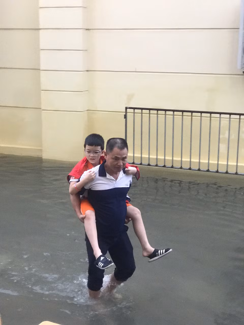 Trước tình hình mưa bão, Trường Tiểu học Nam Trung Yên (Hà Nội) thông báo phụ huynh có thể đón con sớm. Tuy nhiên, mưa lớn kéo dài đã khiến toàn bộ sân trường bị ngập. Phụ huynh phải lội nước cõng con. (Ảnh: PHCC)