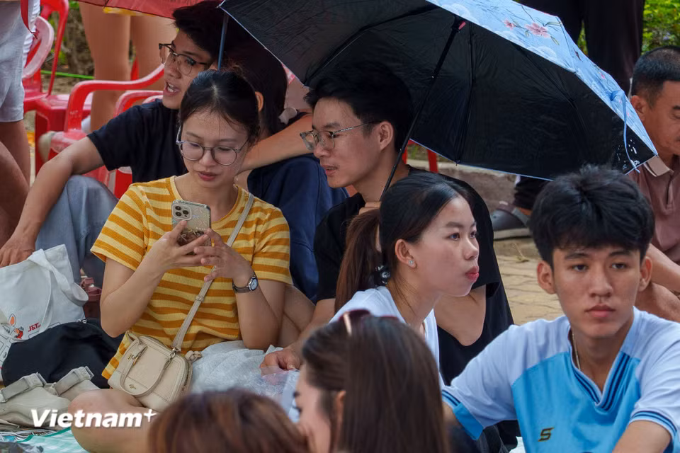 Các bạn trẻ mang theo ô dù, tranh thủ nghỉ ngơi, dùng điện thoại giải trí trong thời gian chờ đợi. (Ảnh: Thanh Phong/Vietnam+)