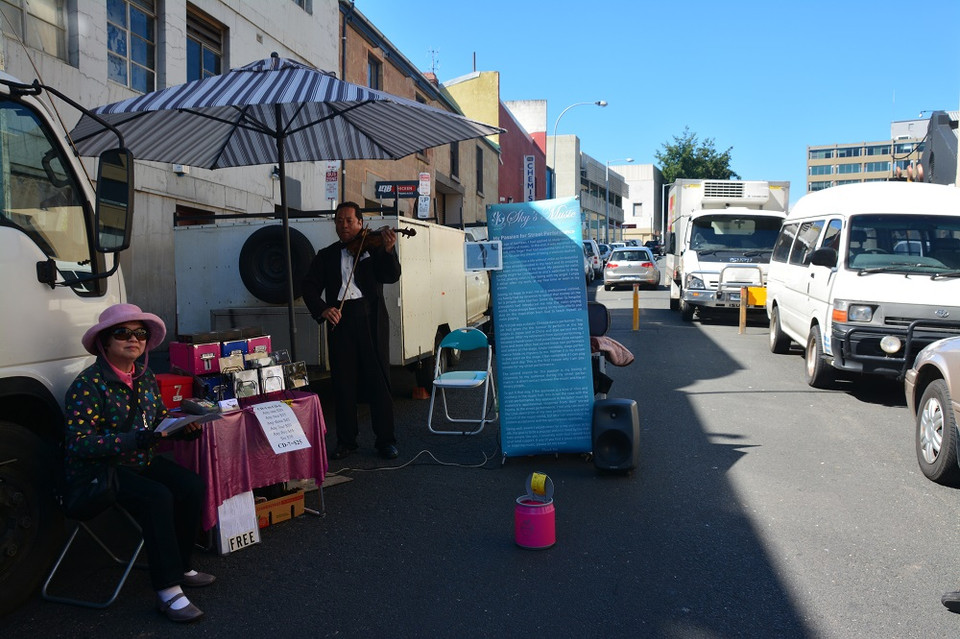 Một “nghệ sỹ” violin châu Á miệt mài biểu diễn bên dòng xe qua lại. Vợ ông chăm chú lắng nghe, không quên giới thiệu với du khách những đĩa CD do chính chồng mình biểu diễn và thu âm. (Ảnh: Võ Giang/Vietnam+)