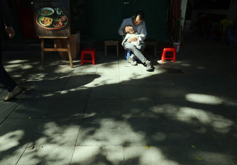 Một người đàn ông cho con trai nhỏ tắm nắng trên vỉa hè phố Lê Thánh Tông. (Ảnh: Lâm Khánh/TTXVN)