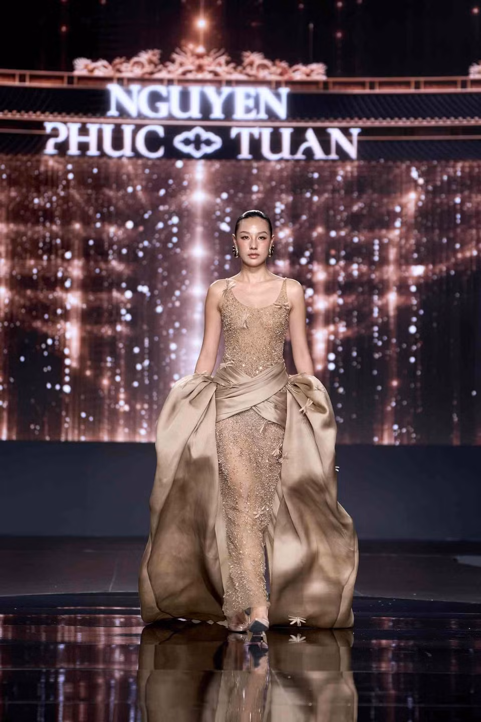 Bảng kỹ thuật trải rộng: dựng phom 3D, xếp lớp, nhún vải, cắt chéo, voan lưới cắt tay, lông vũ nhuộm và tạo hình thủ công, vẽ sơn dầu trên lụa. Ở khâu đính kết, Nguyễn Phúc Tuấn áp dụng kỹ thuật do chính anh phát triển: hạt pha lê được nối vào khung kẽm không gỉ thành từng mảng, cho phép những chuỗi pha lê vươn khỏi mặt vải, rủ dài và rung lên theo từng bước chân như mưa thật. Nhiều thiết kế được làm lại hai đến ba lần cho đến khi đạt đúng cảm xúc. (Ảnh: CTV/Vietnam+)