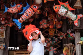 Em nhỏ thích thú đội đầu lân giữa không gian rực rỡ của đèn cá chép, mặt nạ và đồ chơi Trung thu truyền thống. (Ảnh: Phạm Tuấn Anh/TTXVN)